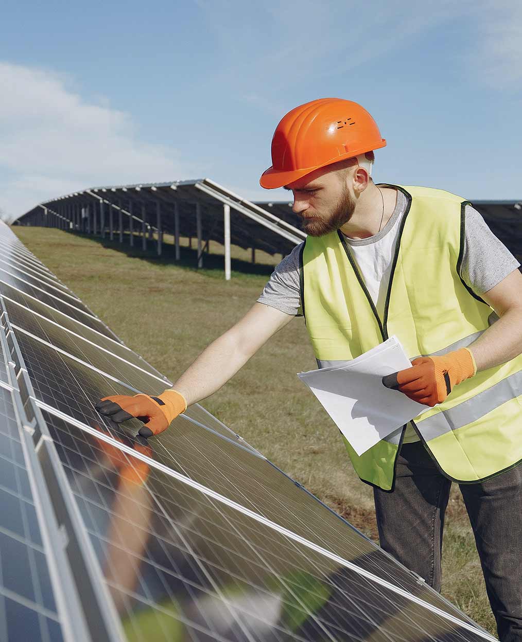 Reduce Costs With Renewable Energy A man with a hard hat and safety vest inspecting solar panels with many panels in the background.